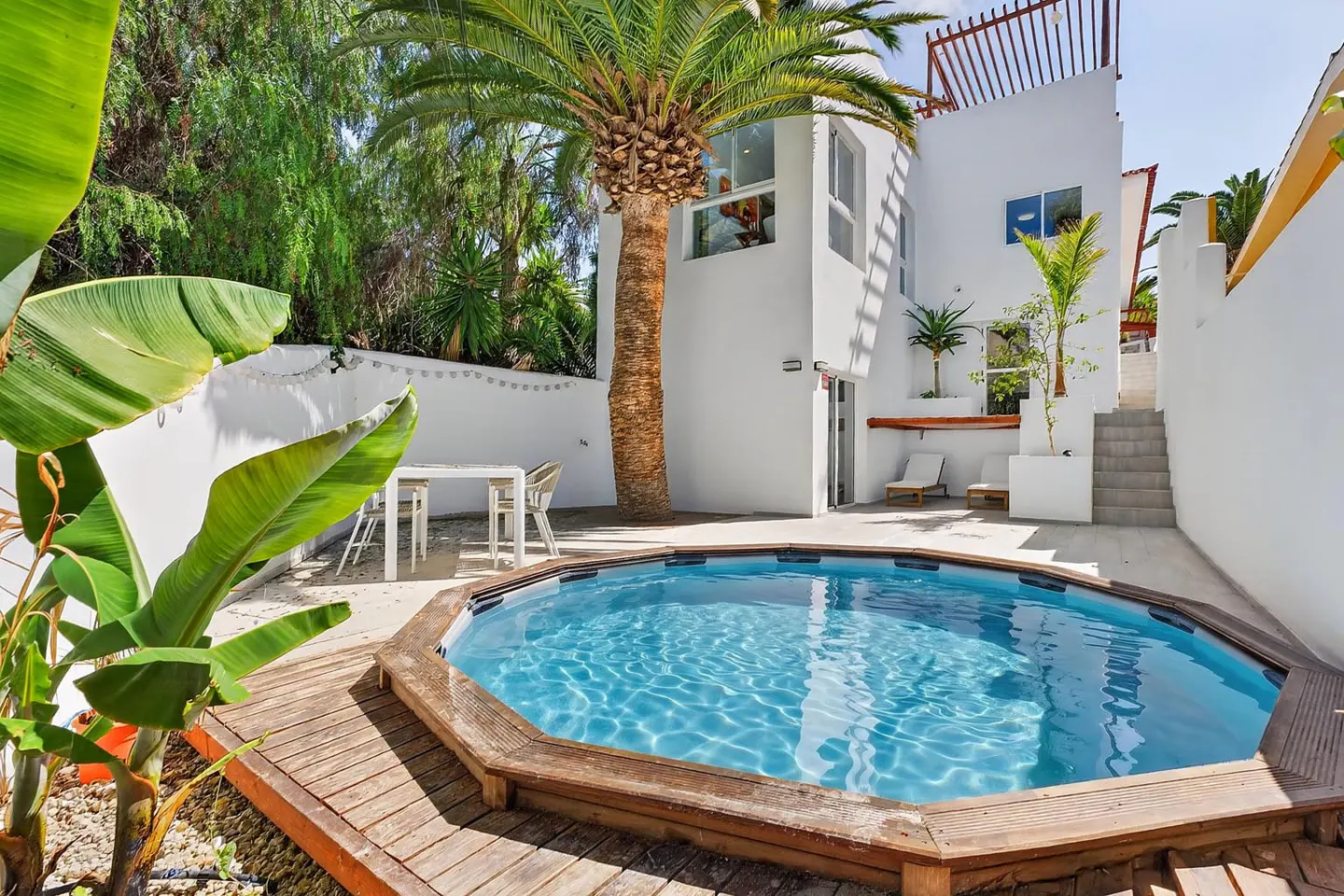 A sunny patio with a circular above-ground pool, wooden decking, a dining set, and a white house with a palm tree.