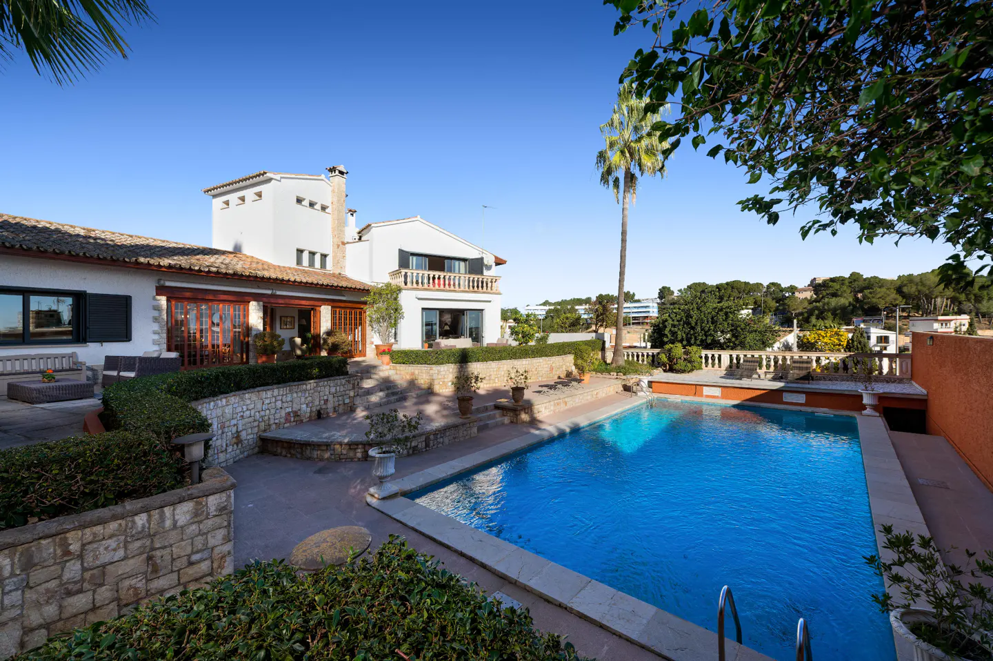 A white house with a red tile roof overlooks a blue swimming pool on a sunny day.