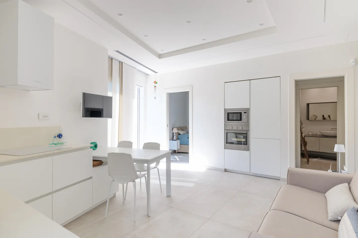 Bright, modern apartment interior with white kitchen, dining table, and beige sofa. Appliances are stainless steel. A bedroom and bathroom are visible.