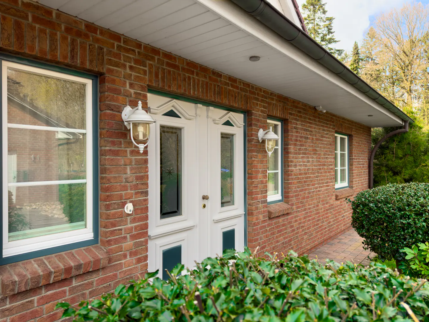 Brick house exterior with white door, windows, and trim. Green bushes in the foreground. Two white sconces flank the door.