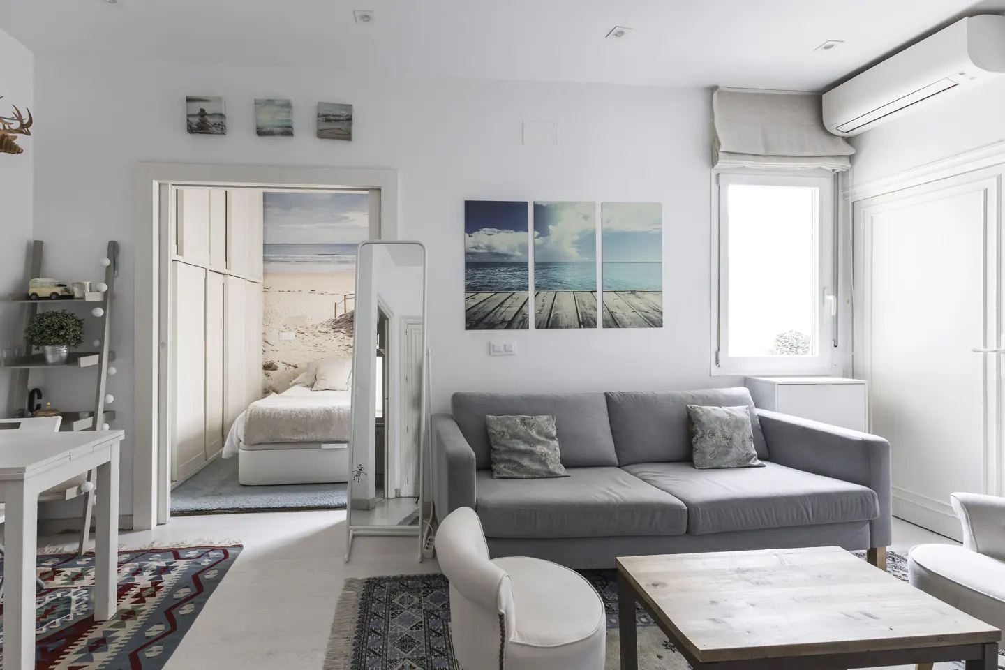 Bright living room with a gray sofa, white walls, and beach-themed art. A bedroom is visible through an open doorway.