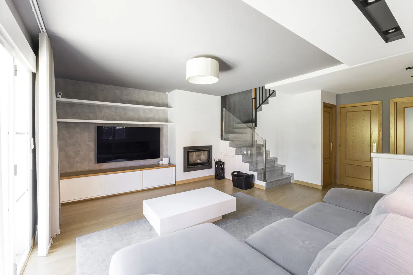 Bright living room with gray sofa, white coffee table, TV, fireplace, and stairs with glass railing.