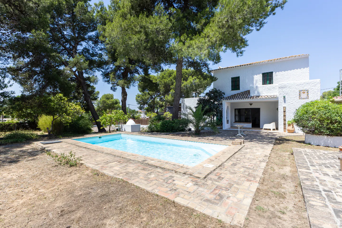 White two-story villa with a swimming pool, brick patio, and large pine trees in a sunny garden setting.