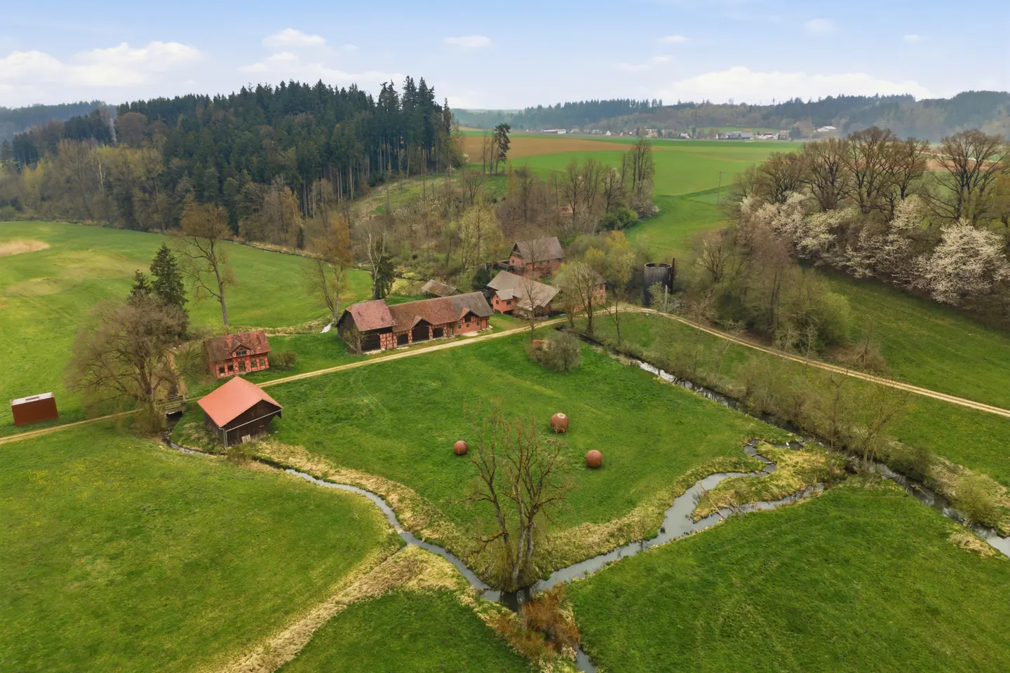Aerial view of a farm with red-roofed buildings, green fields, a stream, and a forest in the background.