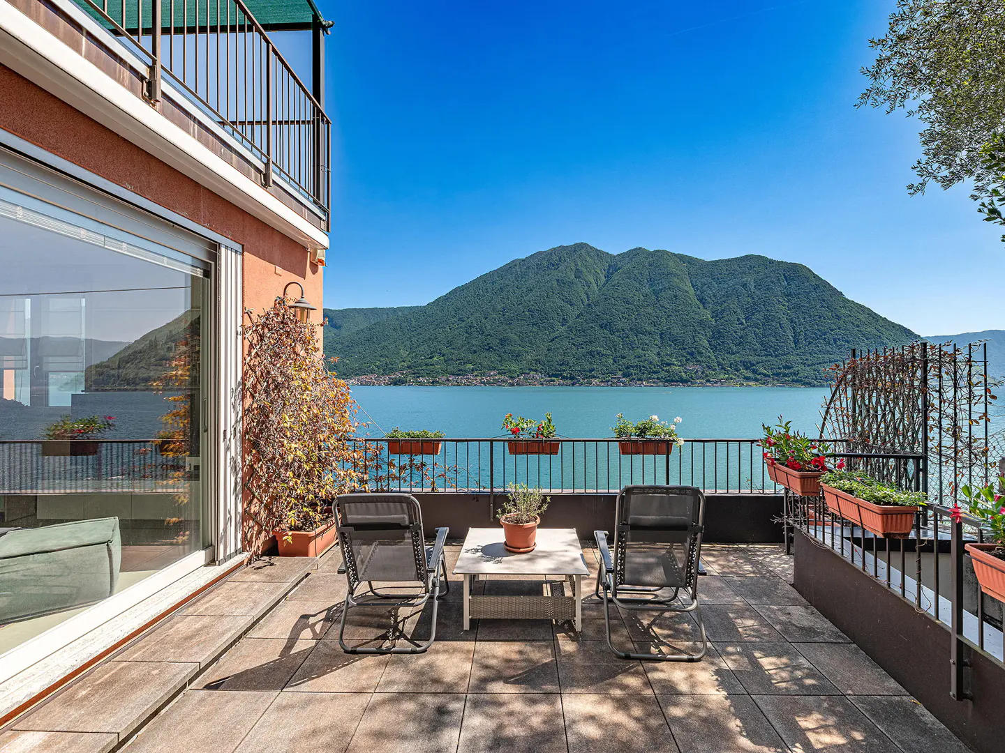 Balcony with two chairs and a table overlooking a lake and mountain on a sunny day.