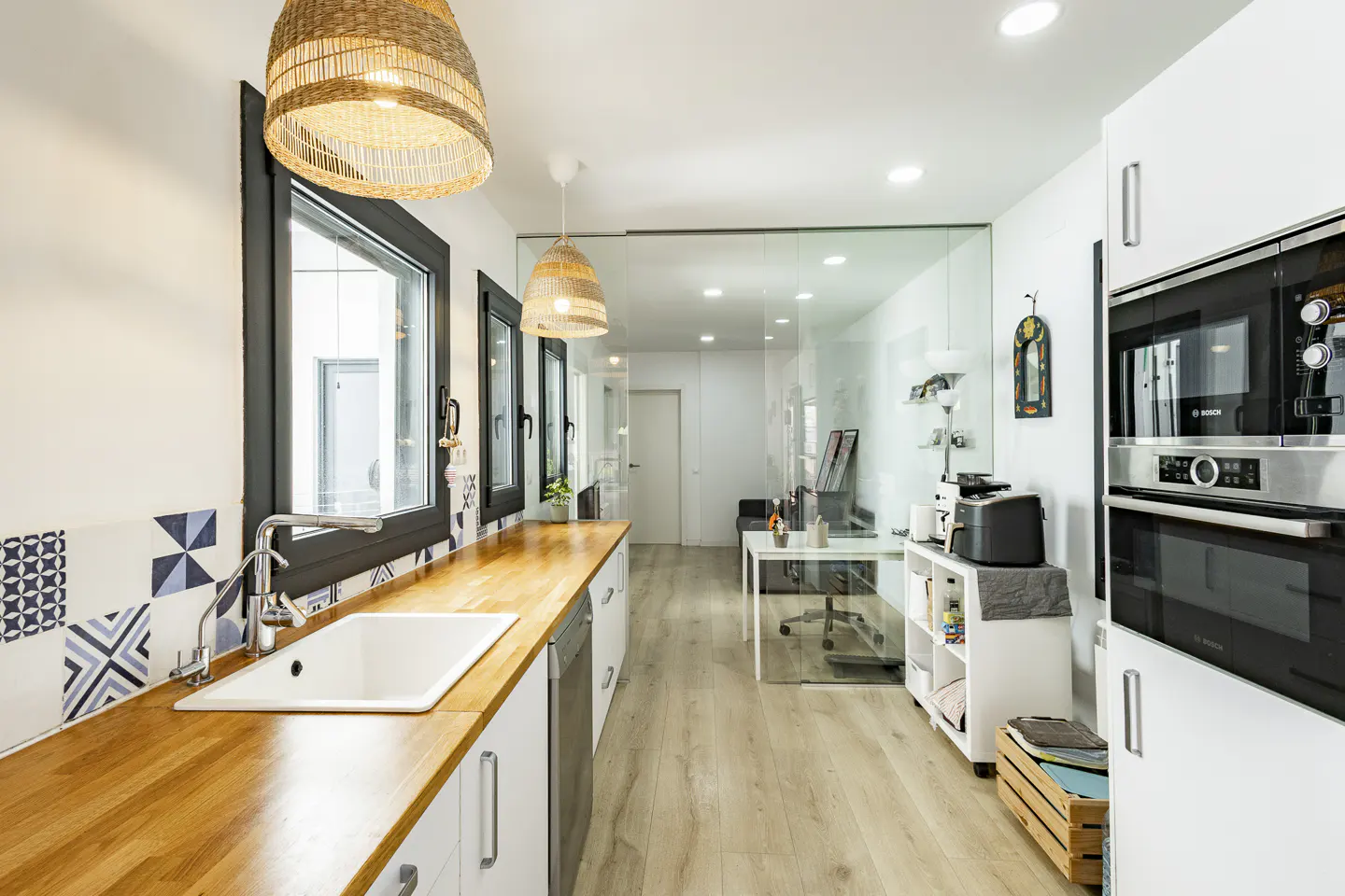 Bright, modern kitchen with wood countertops, white cabinets, and woven pendant lights. A glass-walled office is visible in the background.