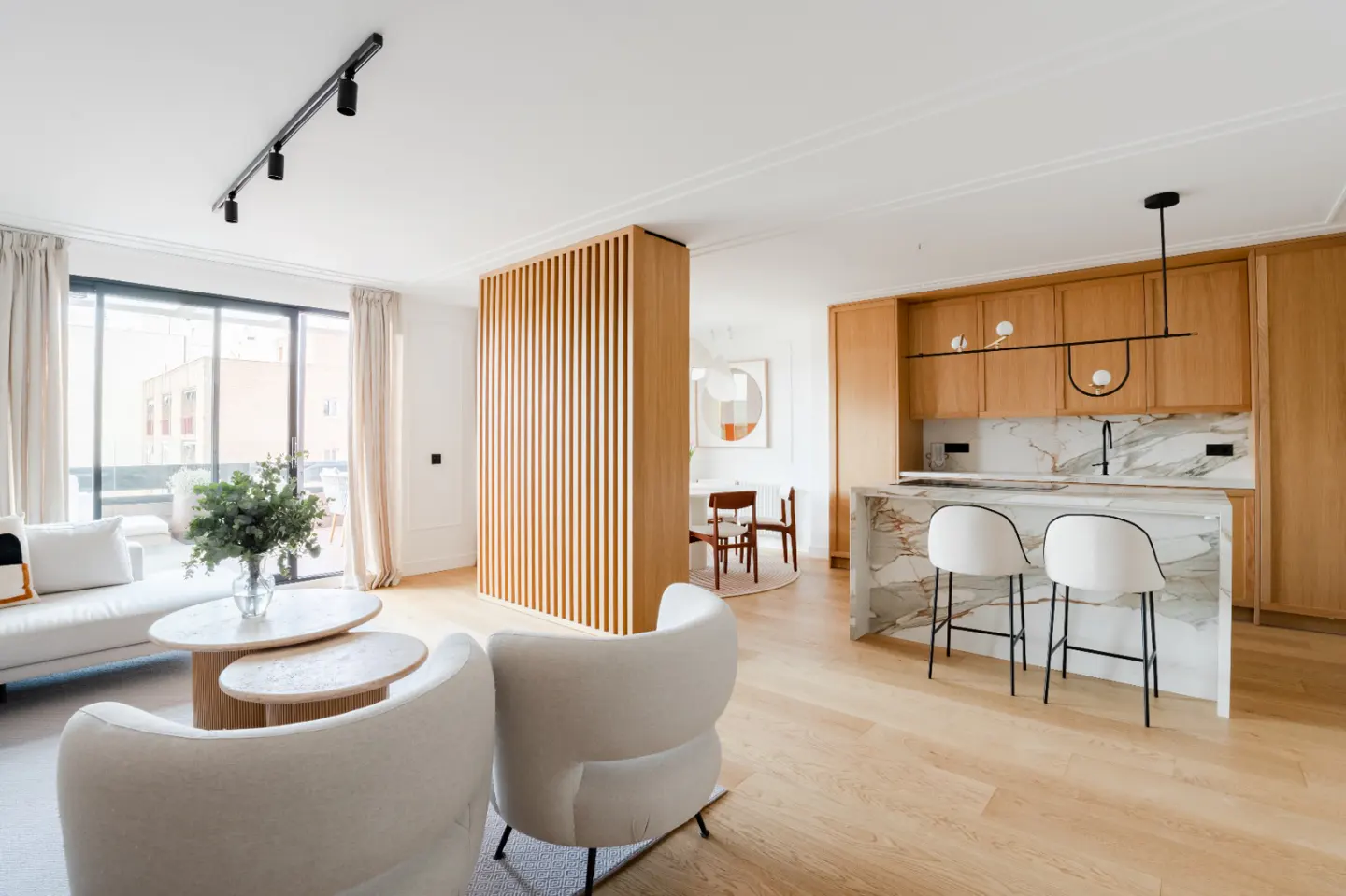 Bright, modern apartment interior with white sofa, chairs, wood floors, and a kitchen island with marble countertop and two white stools.
