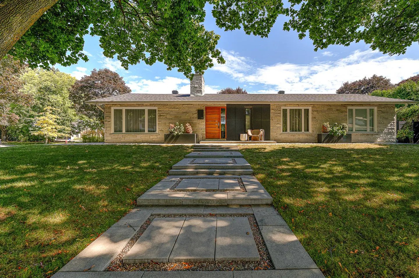 A tan brick house with a stone walkway leading to a wooden door, green lawn, and trees.