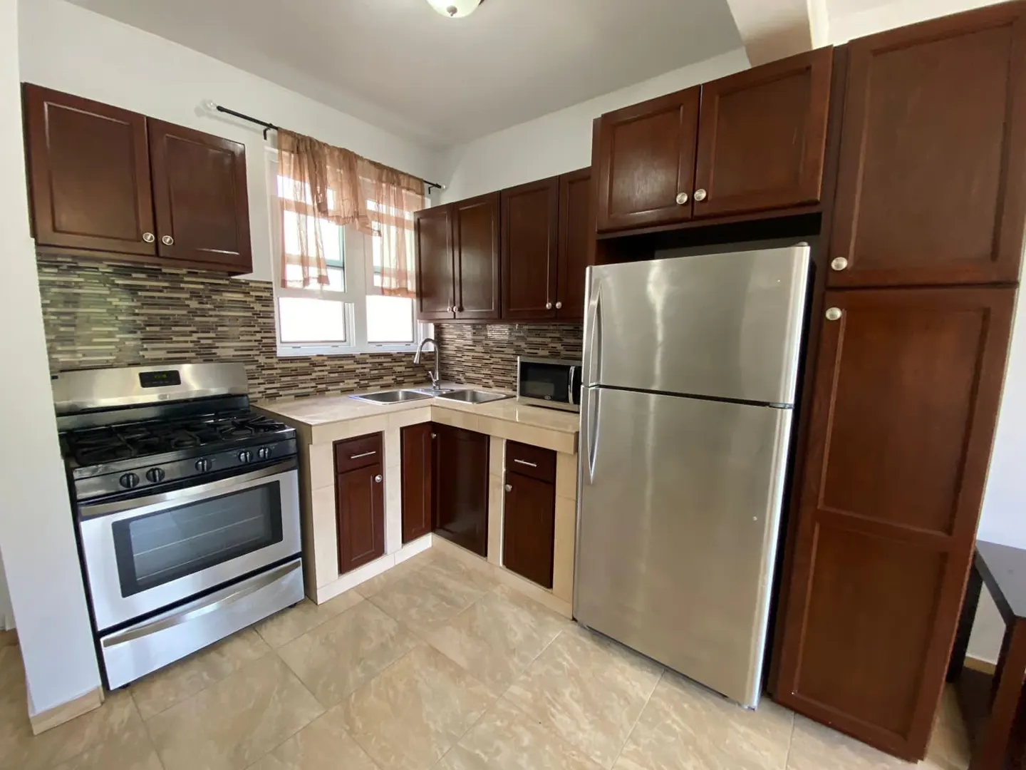 A kitchen with brown cabinets, stainless steel appliances, and beige tile flooring. A window with a brown curtain is above the sink.