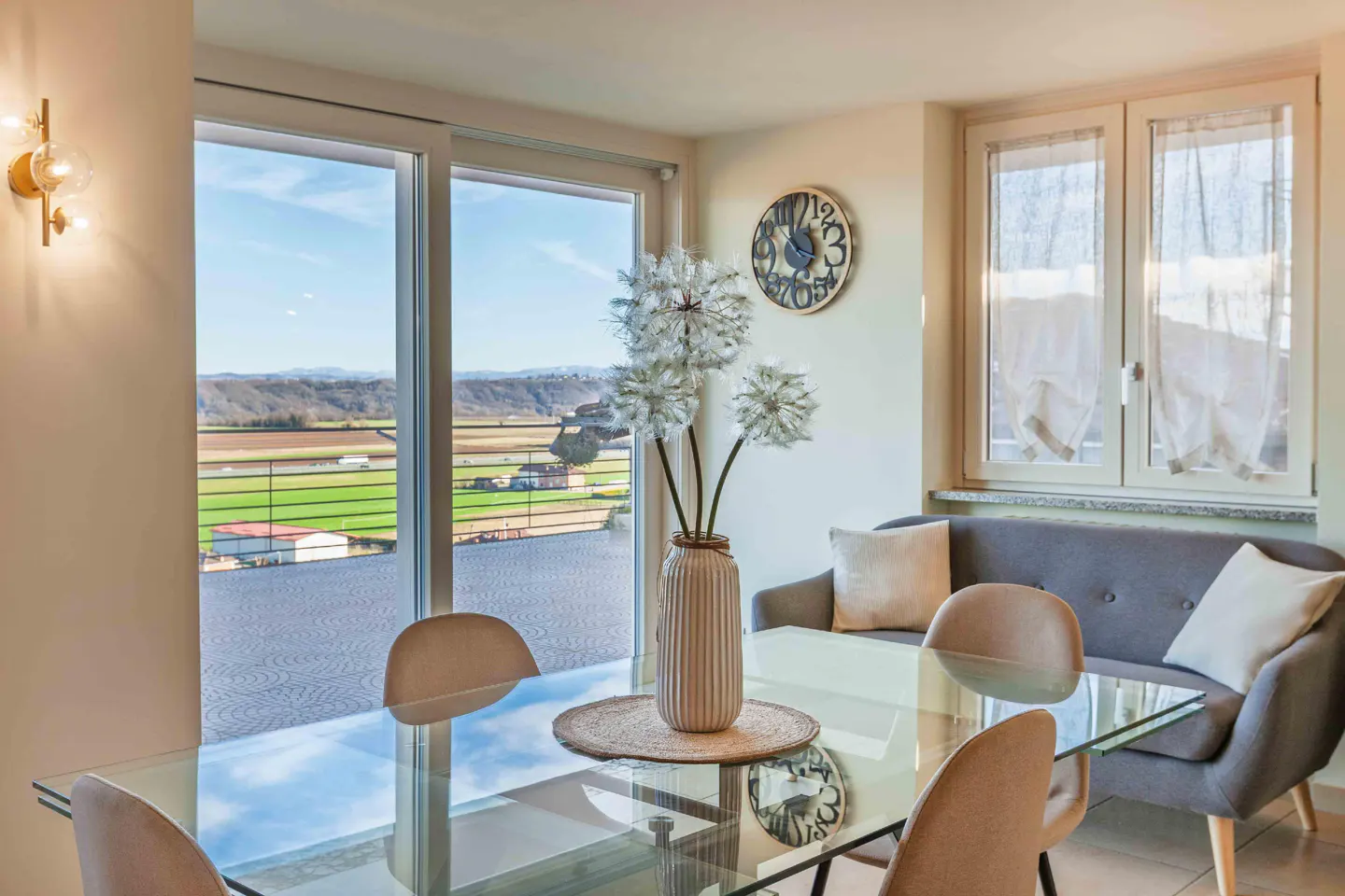 A bright dining area with a glass table, tan chairs, and a gray sofa. A vase of white flowers sits on the table, with a view of green fields through the window.