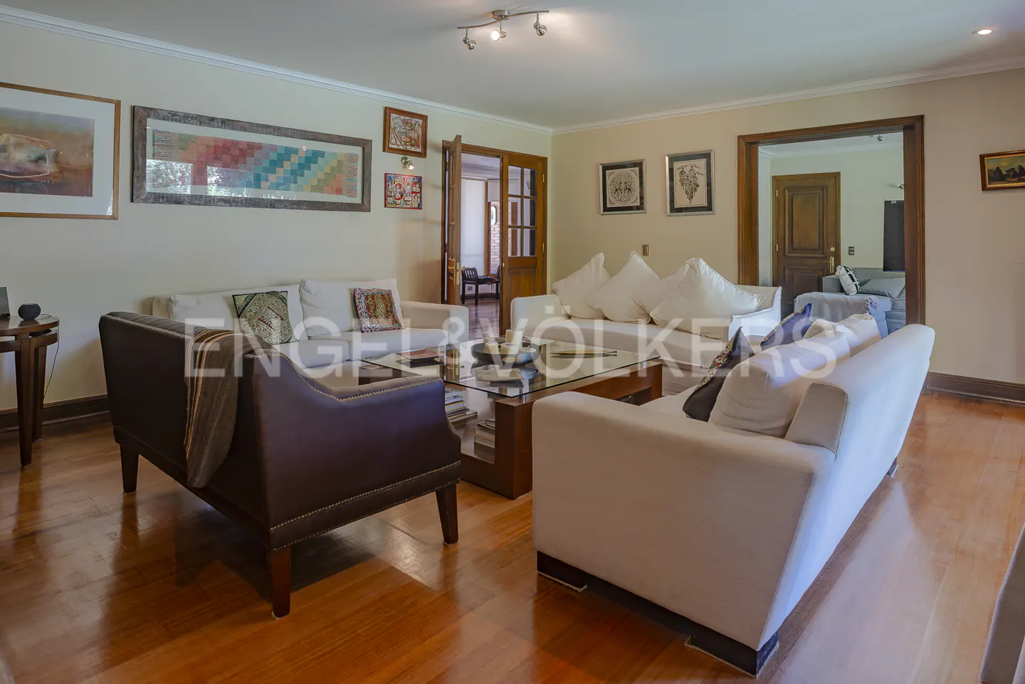 A living room with white sofas, a brown leather chair, and a glass coffee table on a wooden floor. Artwork hangs on the walls.