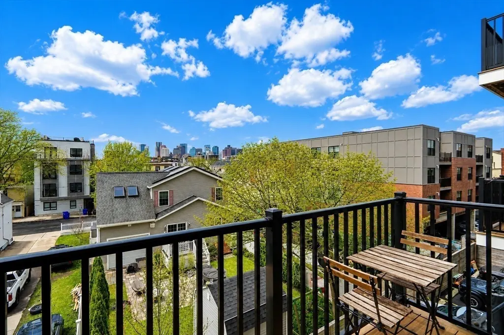 Balcony view with a small wood table and chairs, black railing, houses, trees, and a city skyline under a blue sky with white clouds.