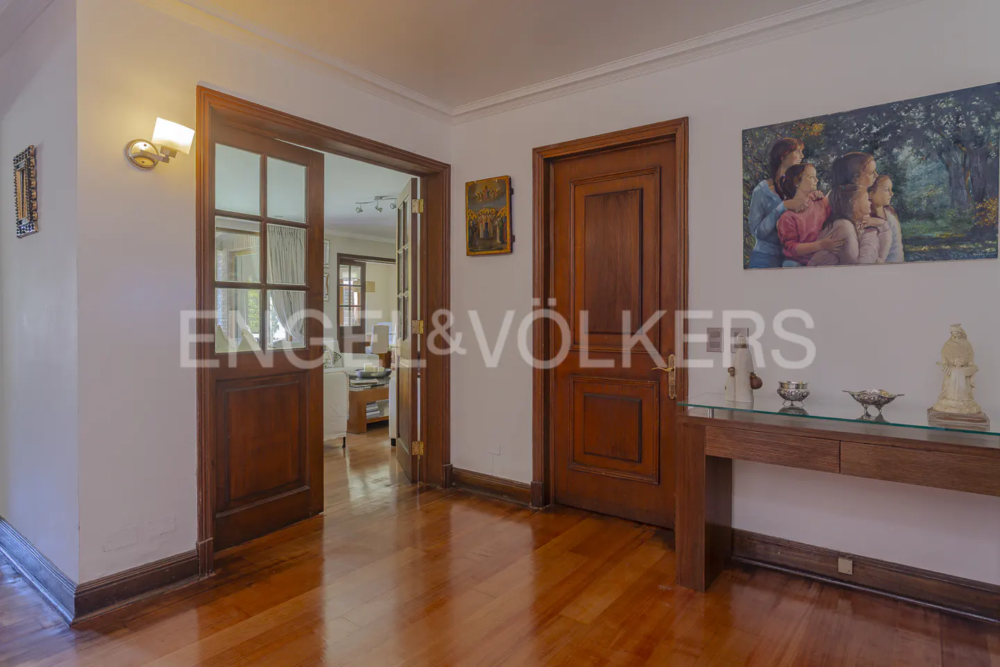 A foyer with hardwood floors, white walls, and dark wood trim. A glass-topped console table holds decorative objects. A painting hangs above a closed wood door.