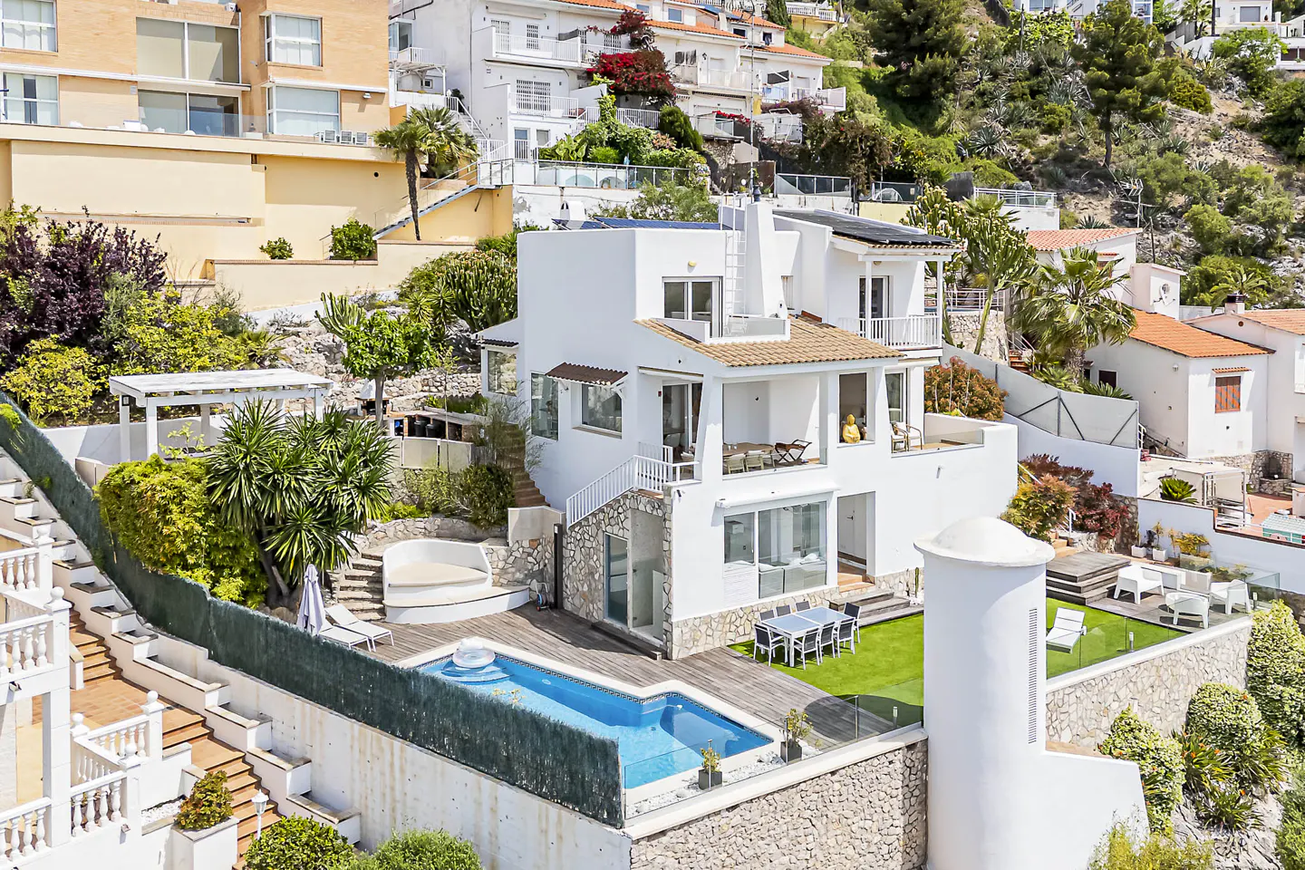Multi-level white villa on a hillside with a pool, patio, and green lawn. Palm trees and other houses are in the background.