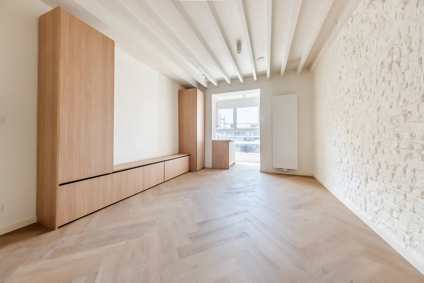 Bright, empty room with herringbone wood floors, white brick wall, and light wood built-in cabinets. White ceiling with exposed beams. Window with outside view.