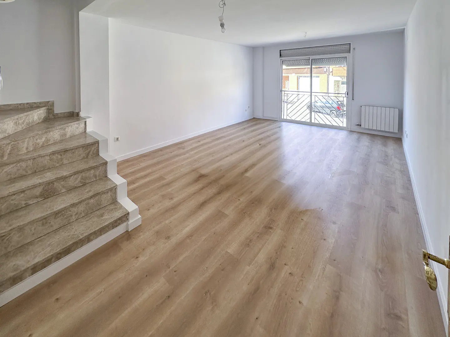 Empty room with light wood floors, white walls, marble-look stairs on the left, and a sliding glass door to a balcony.