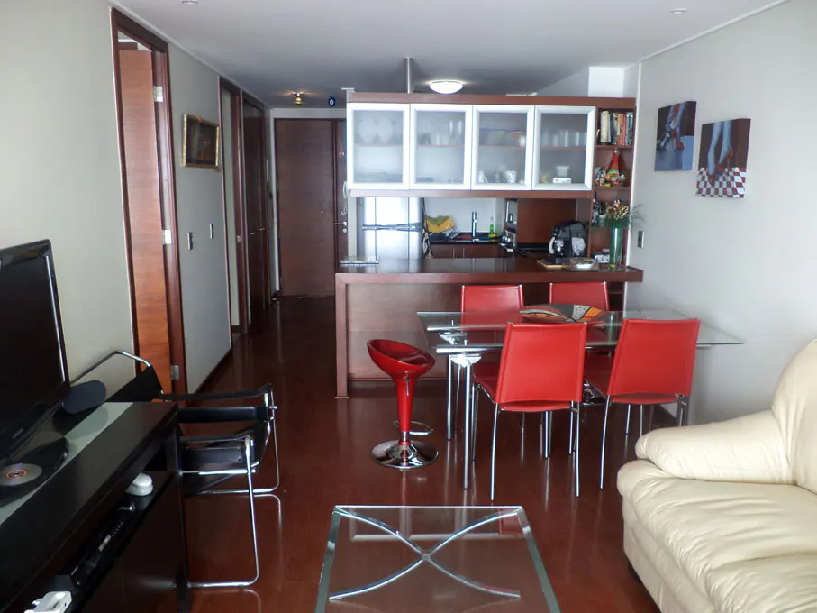Interior view of a modern apartment with a kitchen, dining area, and living room. Red chairs surround a glass table. A white sofa is visible.