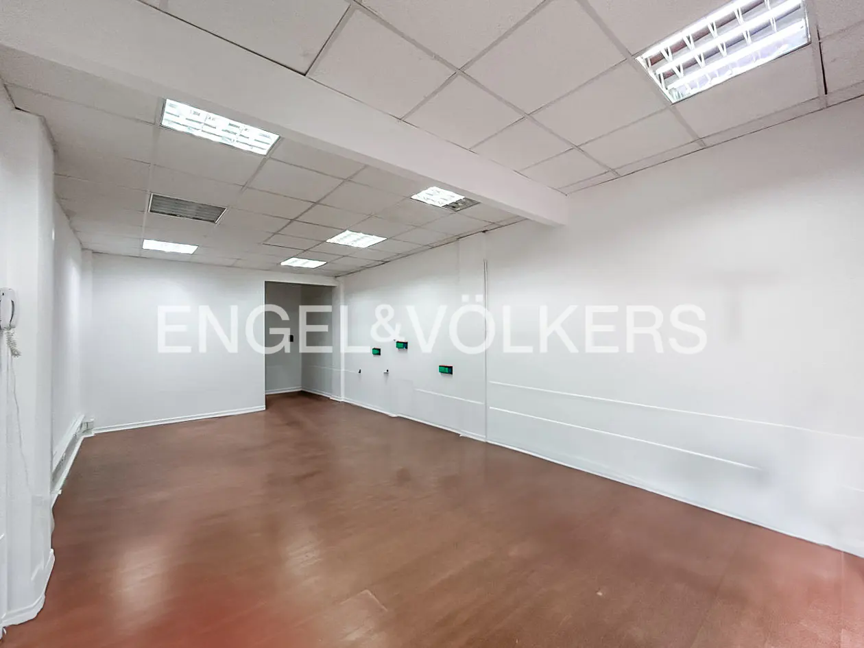 Empty commercial space with white walls, brown floor, and fluorescent lighting. Engel & Völkers logo on the wall.