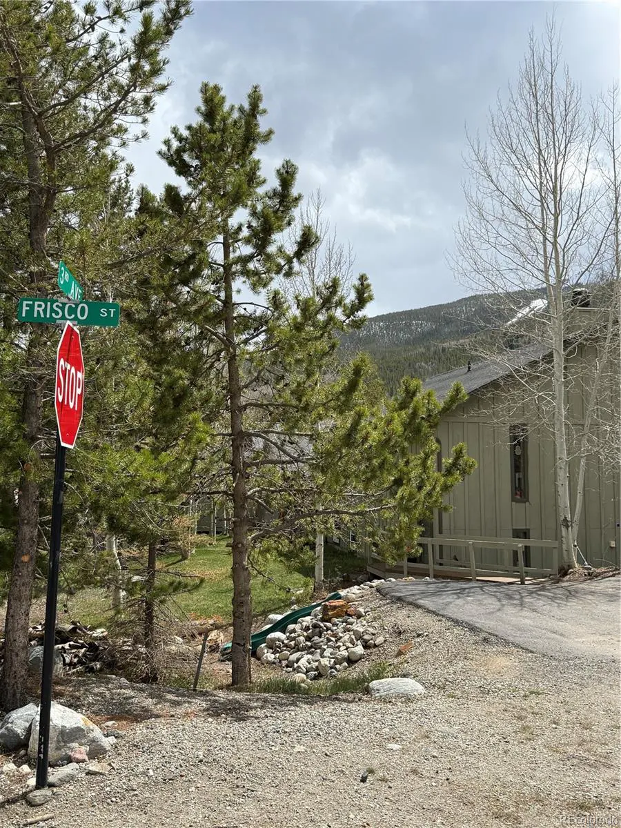 Street view of Frisco, Colorado, with a stop sign, pine trees, and a building in the background. Mountains are visible in the distance.