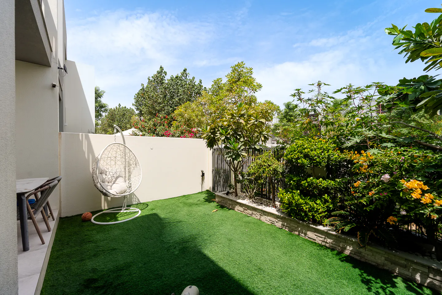 Backyard with green turf, a white egg chair, and lush greenery against a blue sky.