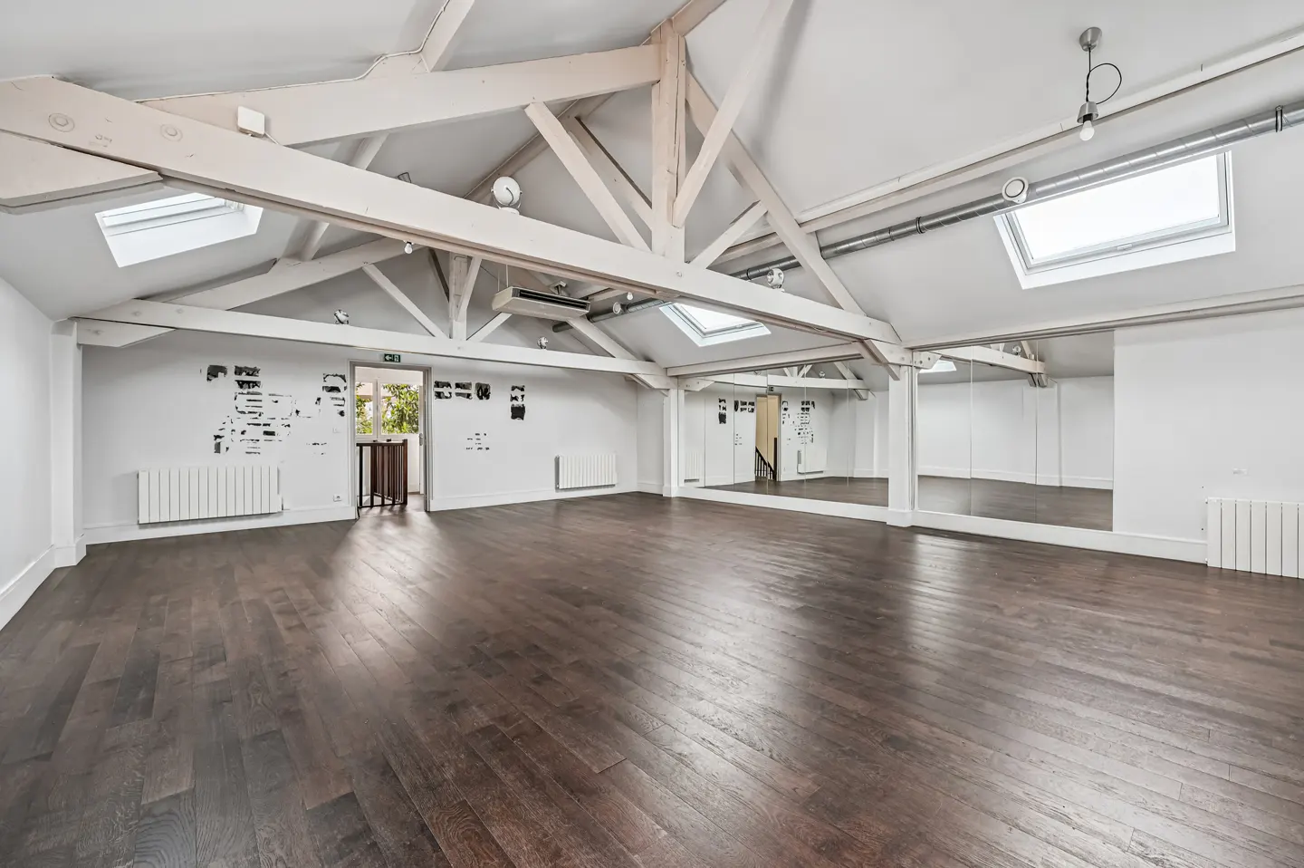 Bright, empty studio with dark wood floors, white walls, and exposed white beams. Mirrors line one wall. Skylights provide natural light.