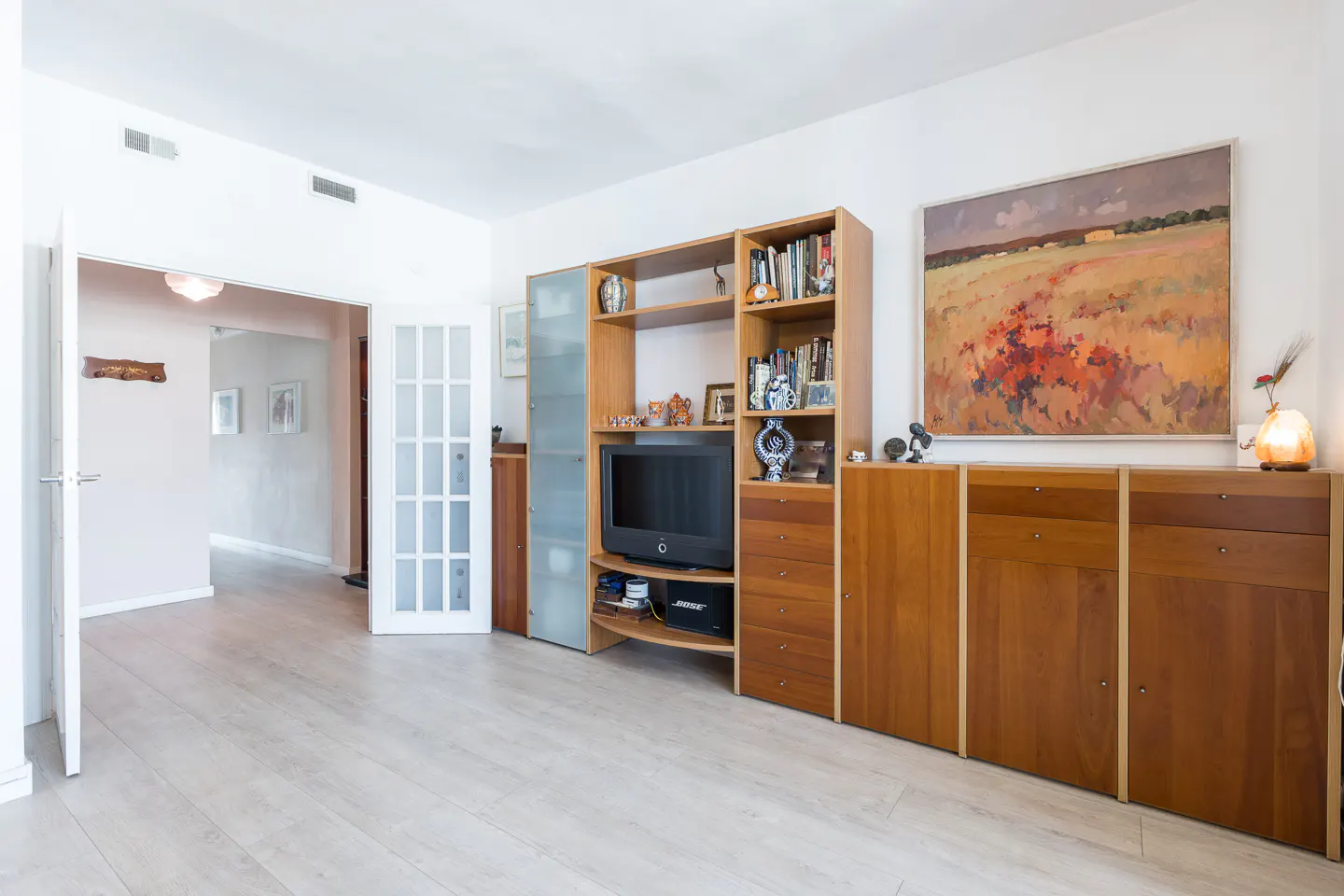 Living room with light wood floors, white walls, and a large wooden entertainment center. A landscape painting hangs above a cabinet. Open doors lead to another room.