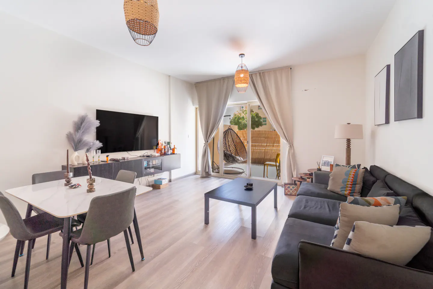 A bright living room with a dining table, TV, and a dark gray sofa. Sliding glass doors lead to a patio with a hanging chair.