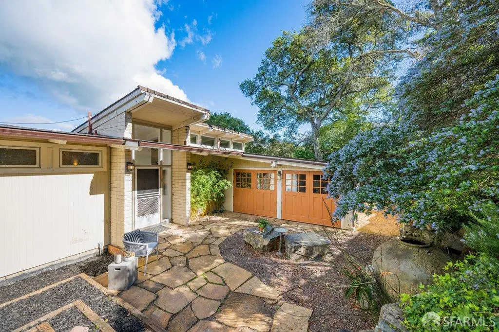 Exterior view of a tan mid-century modern home with orange garage doors, stone patio, and lush greenery under a blue sky.
