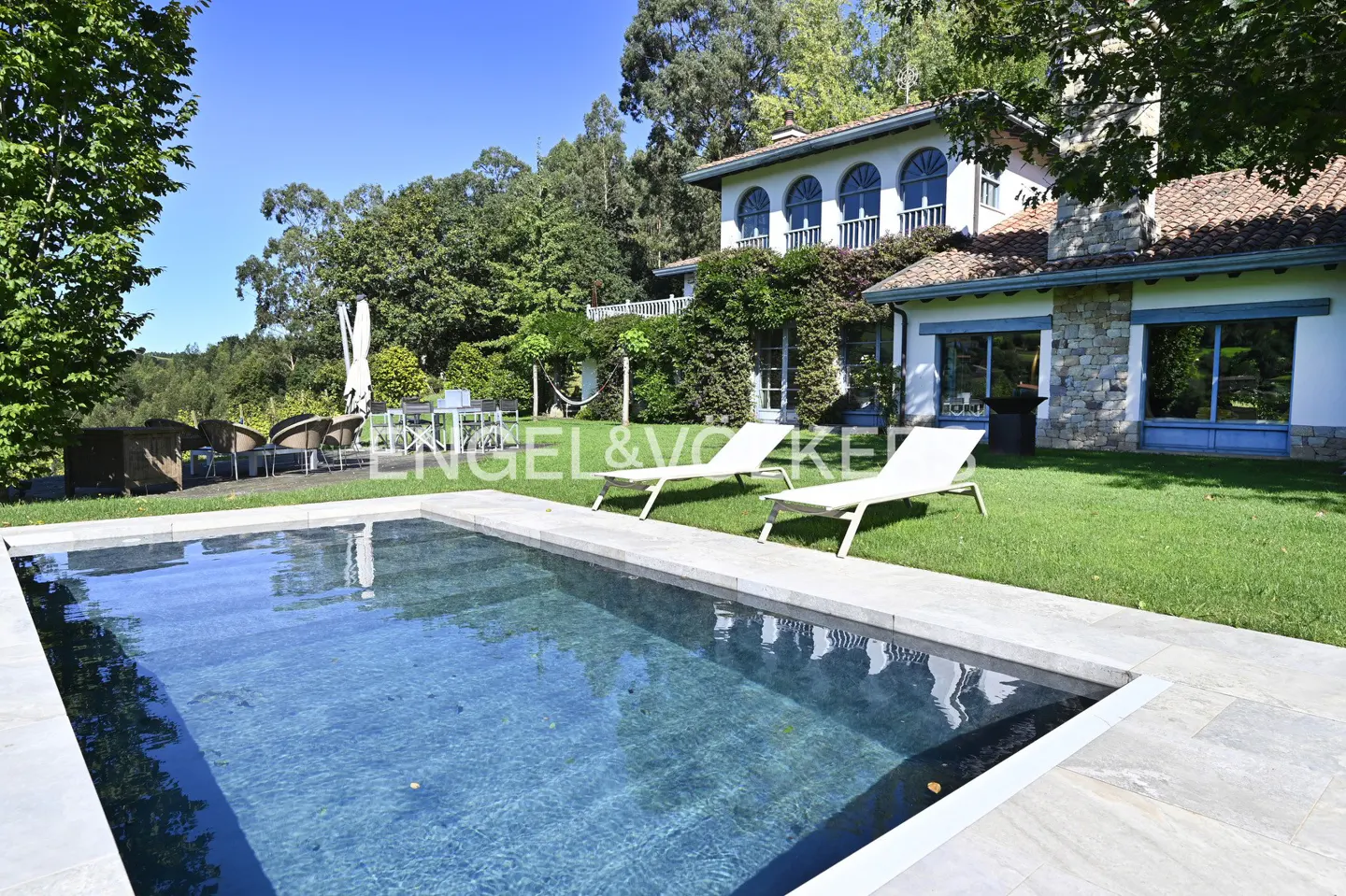 A backyard view of a white house with a pool, two white lounge chairs, and green grass.