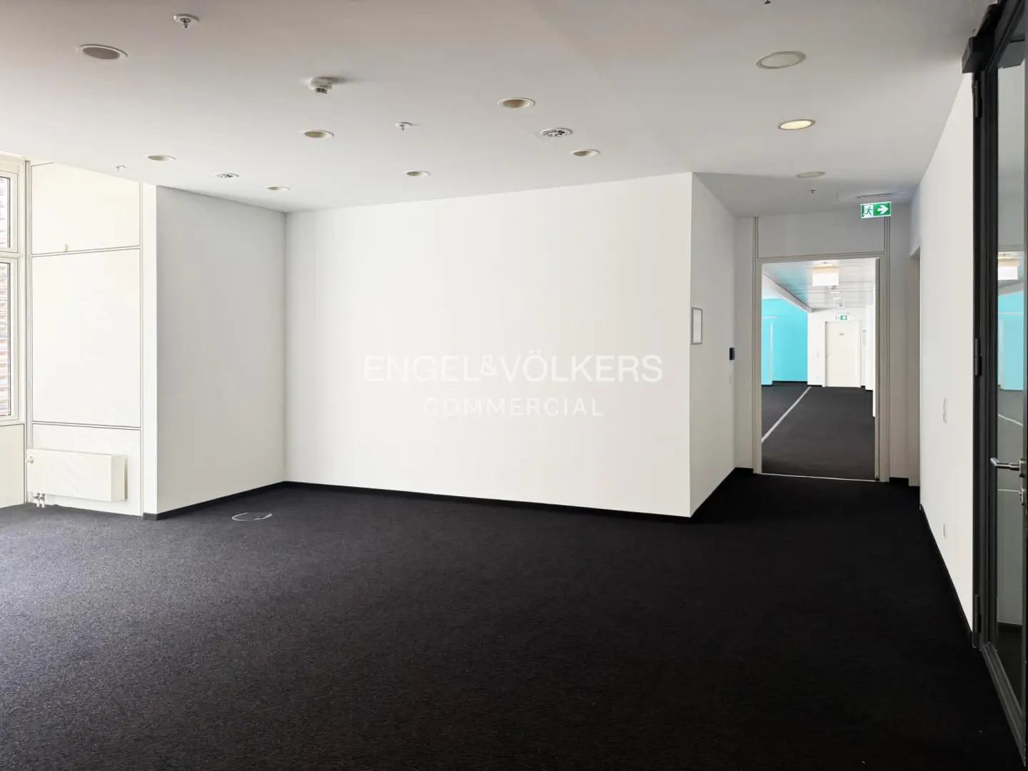 Empty office space with black carpet, white walls, and recessed lighting. Engel & Völkers Commercial logo on the wall. Hallway with exit sign visible.