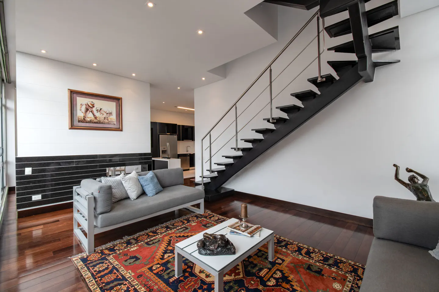 Bright living room with a gray sofa, patterned rug, and black staircase. A painting hangs above a black tiled fireplace.