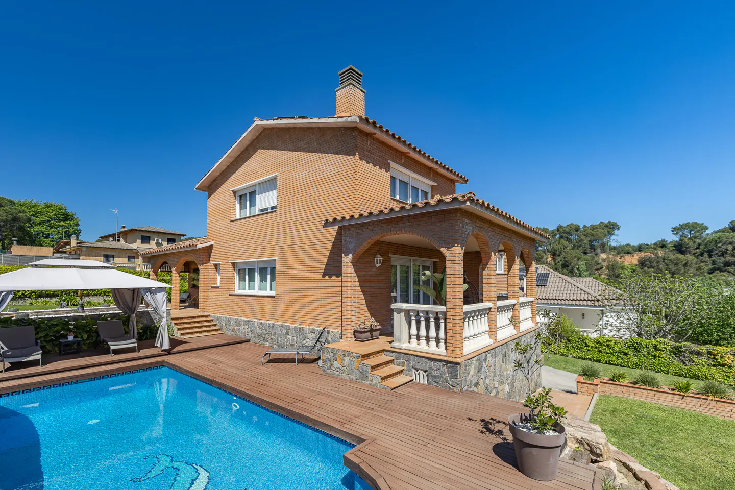 Two-story brick house with a pool and wooden deck under a clear blue sky. A white gazebo sits near the pool.