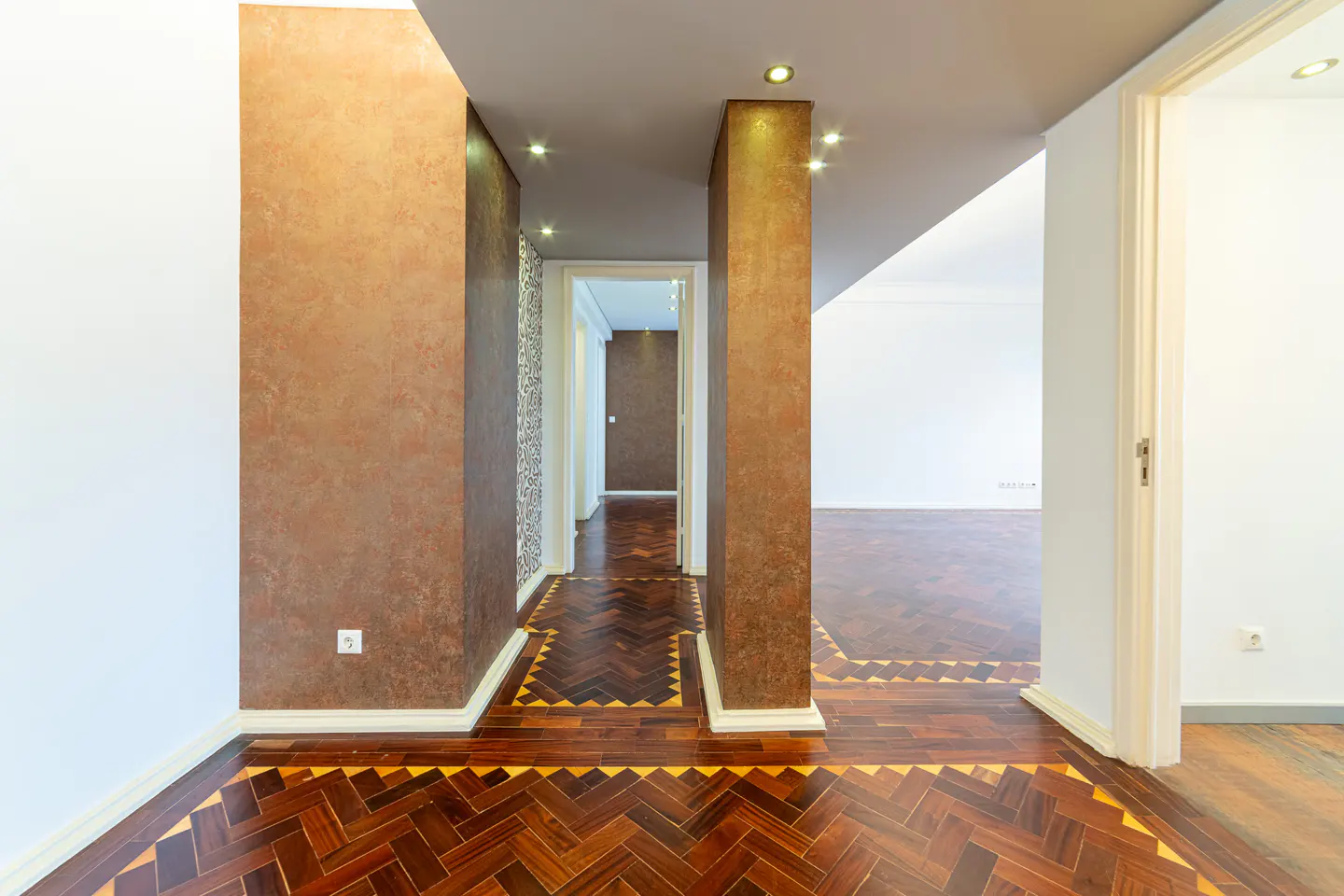 Hallway with herringbone wood floors, brown textured walls, and white trim. Open doorways lead to other rooms.