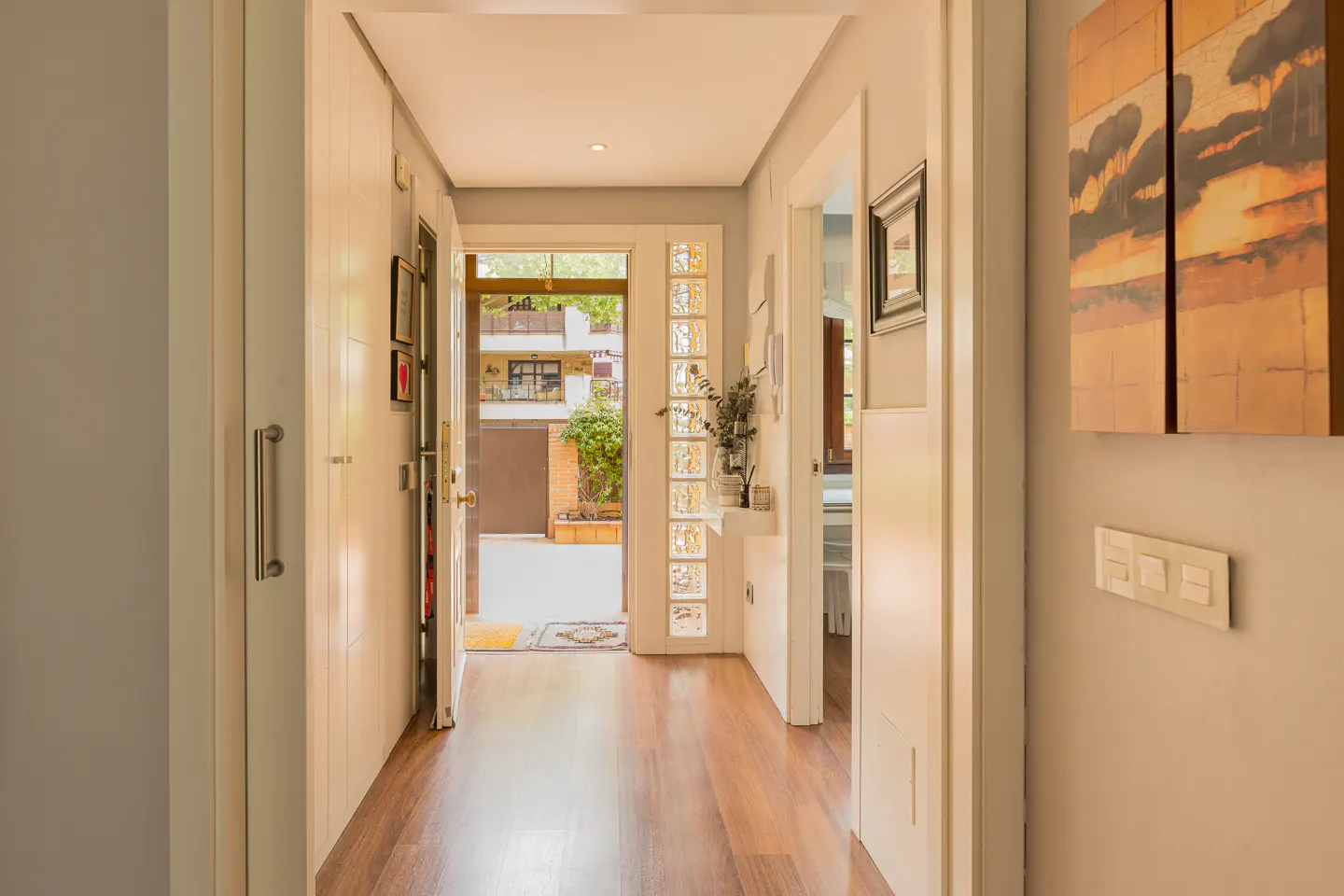 Hallway view with wood floors, white doors, and a glass-block wall near the open front door. Artwork hangs on the wall.