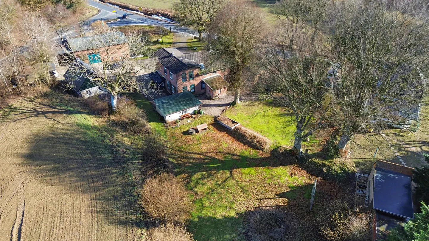 Aerial view of a brick house with green shutters, surrounded by trees and a field. A road is visible in the background.