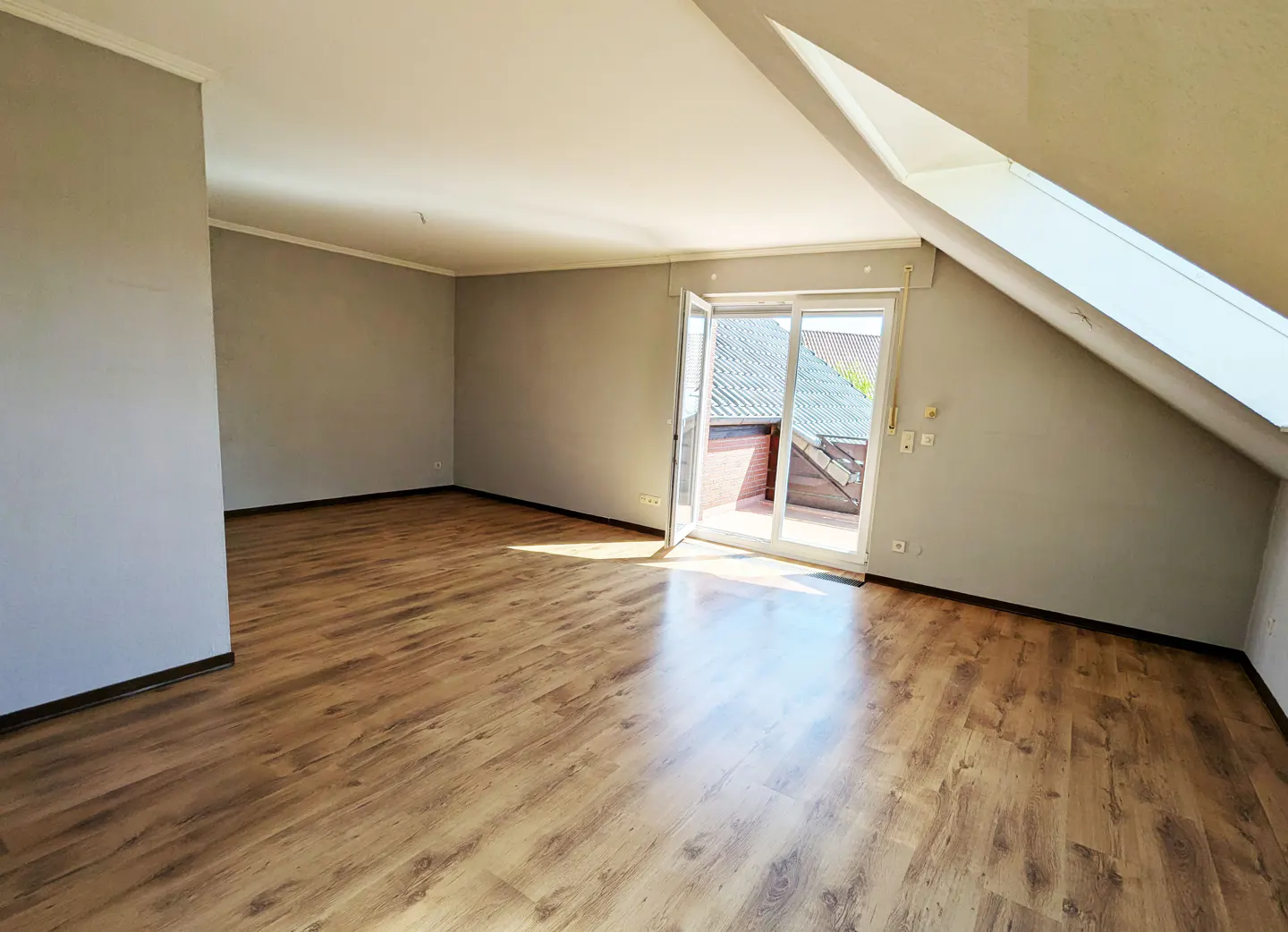 Attic room with wood floor, gray walls, and white trim. Sliding glass doors lead to a balcony. Sunlight streams in.