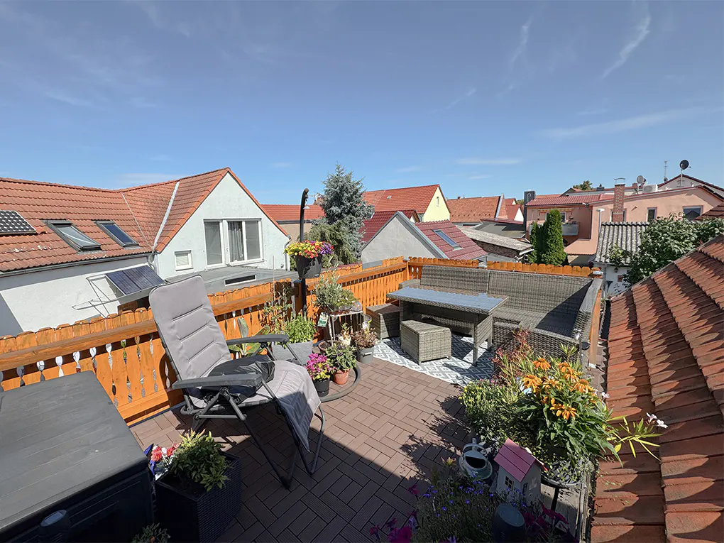 Rooftop patio with gray wicker furniture, a lounge chair, and potted plants. Red-tiled roofs of neighboring houses are visible under a blue sky.