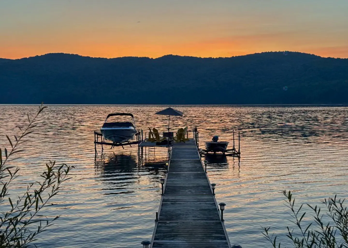 Sunset view of a lake with a wooden dock leading to a boat lift, chairs, and a small boat. Mountains are visible in the background.