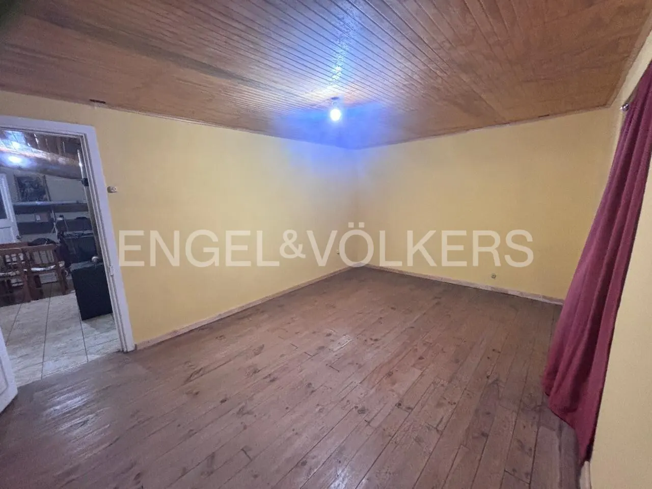 Unfurnished room with wood floors, yellow walls, and wood ceiling. A doorway leads to another room. Engel & Völkers logo on the wall.