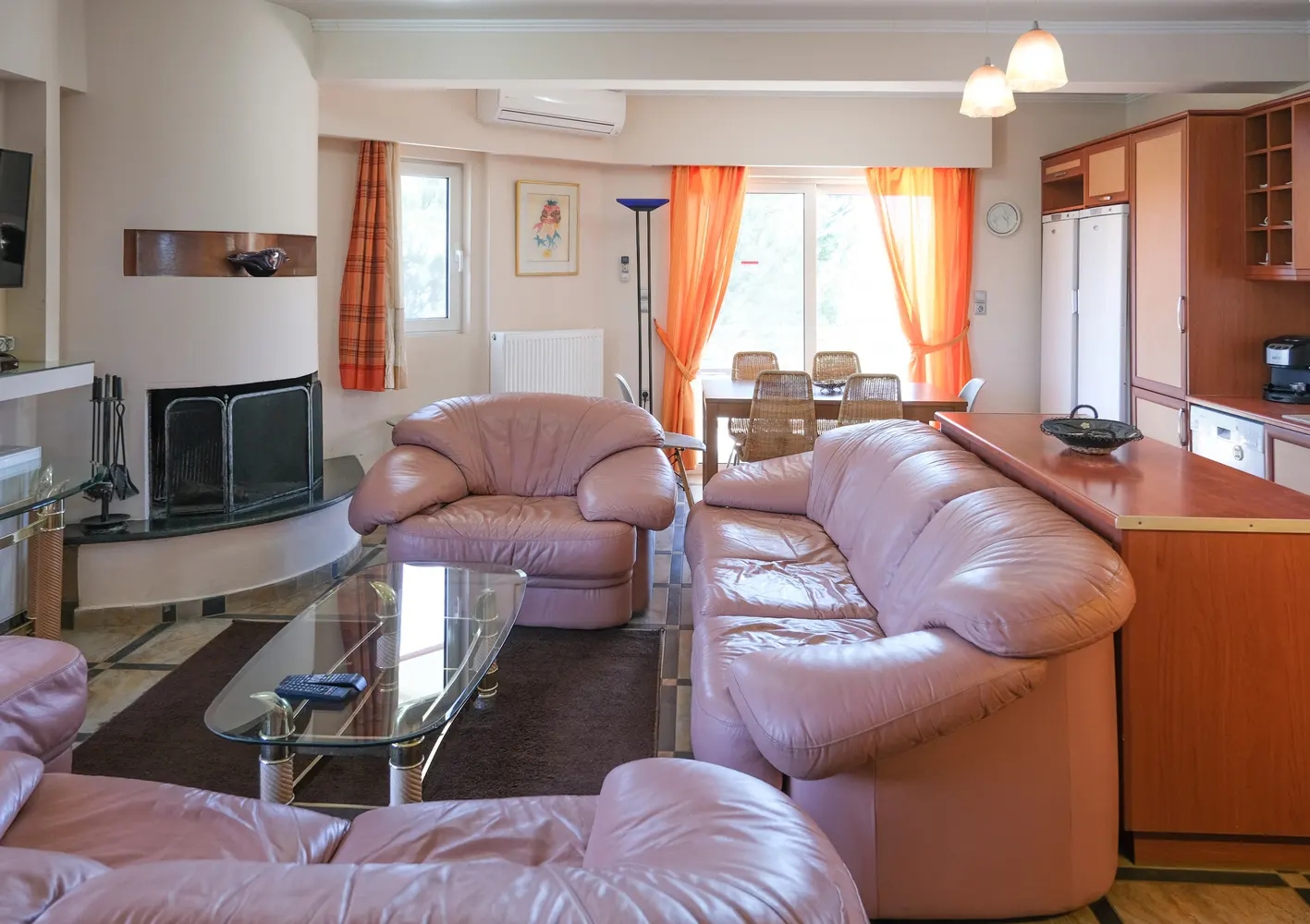 Living room with pink leather sofas, glass table, and fireplace. Orange curtains frame a dining area with wicker chairs and a kitchen with wood cabinets.
