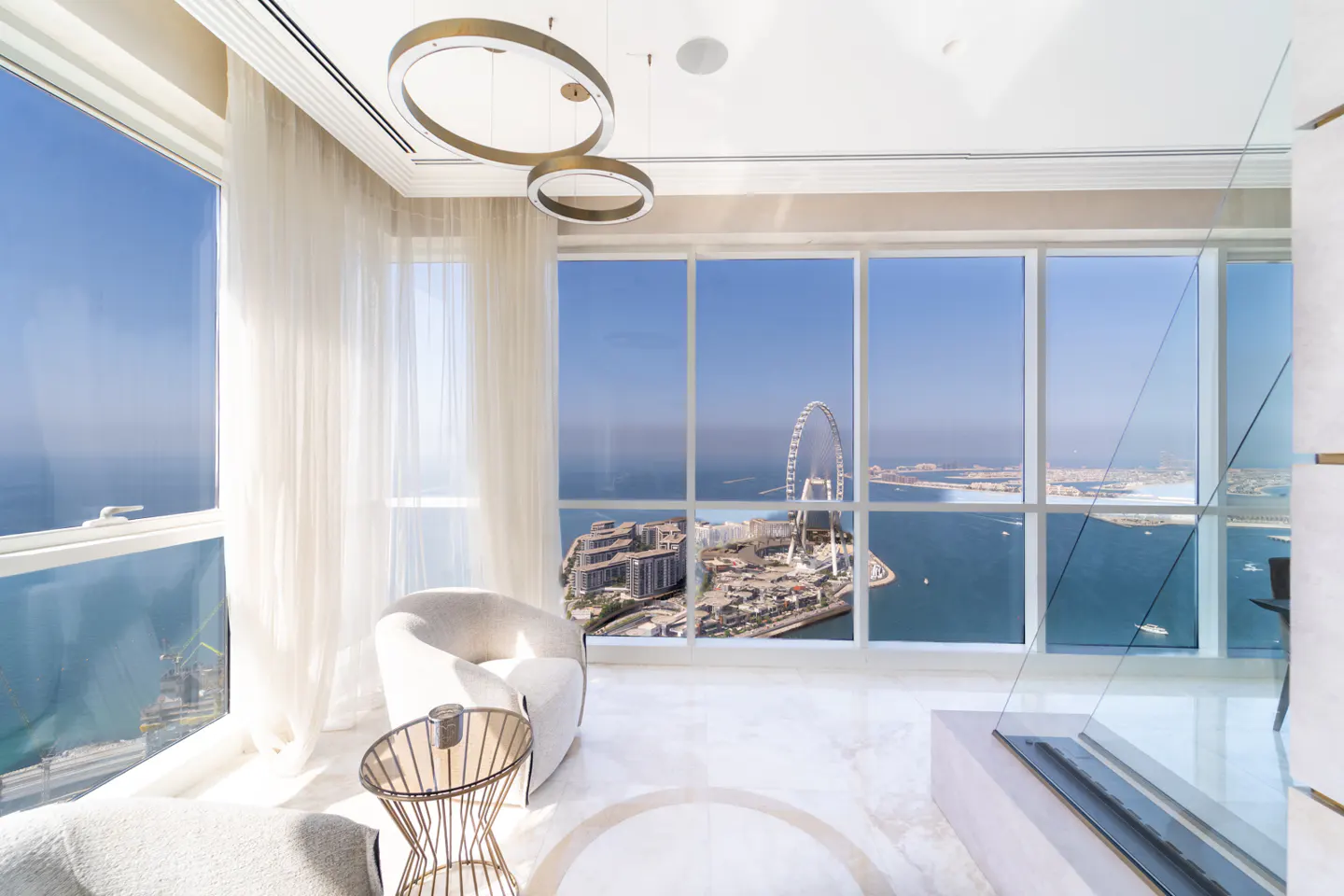 Bright, modern living room with floor-to-ceiling windows overlooking a city skyline and ocean. White chairs, marble floors, and gold accents.