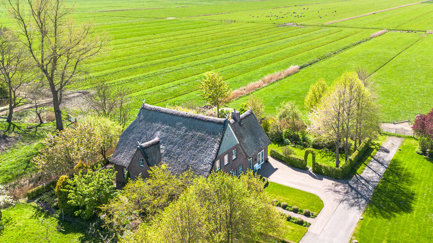 Aerial view of a brown brick house with a thatched roof, surrounded by lush green fields and trees on a sunny day.