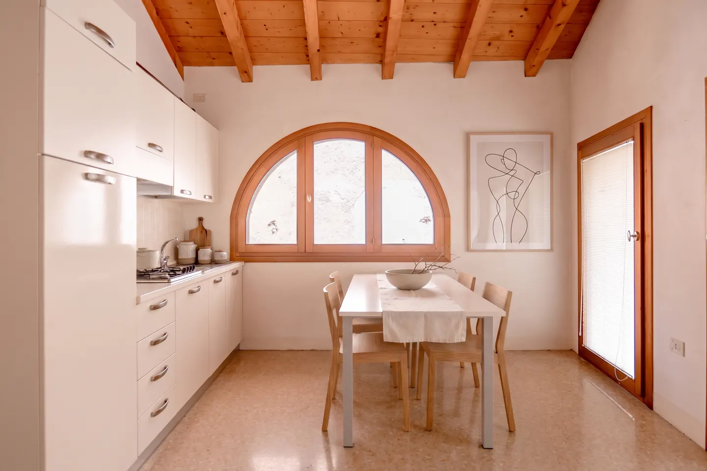 Bright kitchen with white cabinets, wooden ceiling beams, arched window, and a dining table with four chairs.