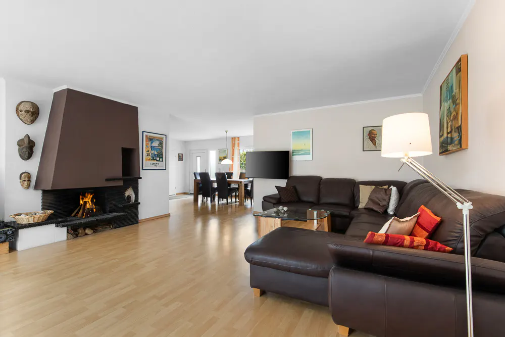 Living room with a brown leather sectional sofa, a fireplace with tribal masks, and a dining area in the background.