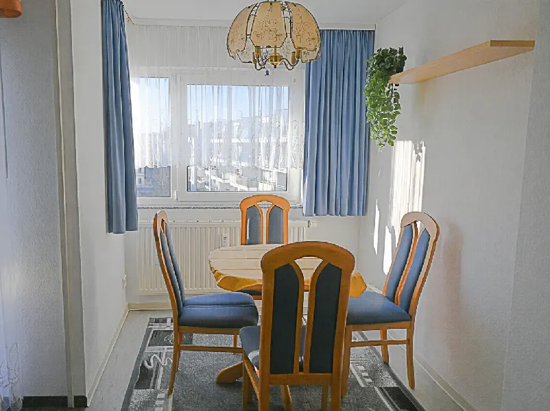Dining room with a round wooden table, four blue chairs, and a patterned chandelier. Blue curtains frame a window with a city view.