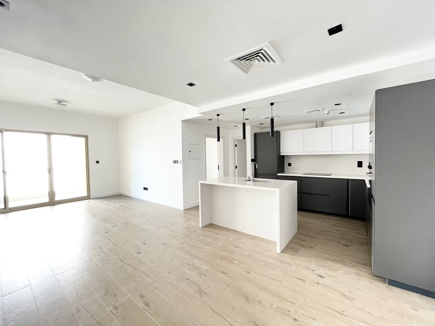 Bright, modern kitchen and living area with light wood floors, white walls, and gray and white cabinets. A white island stands in the center.