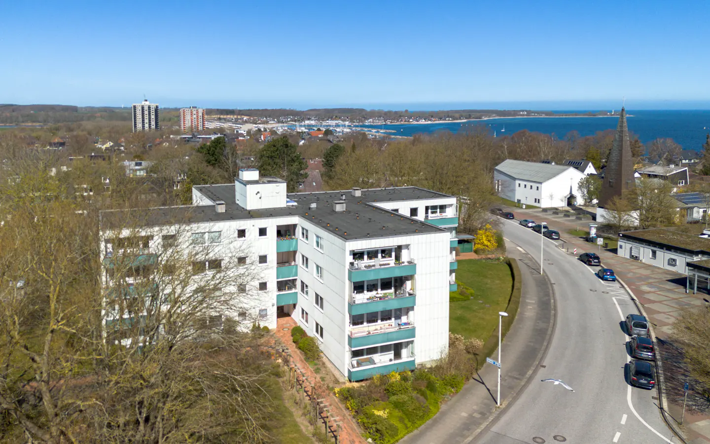 Aerial view of a white apartment building with green balconies, a road, and a church steeple in the background.