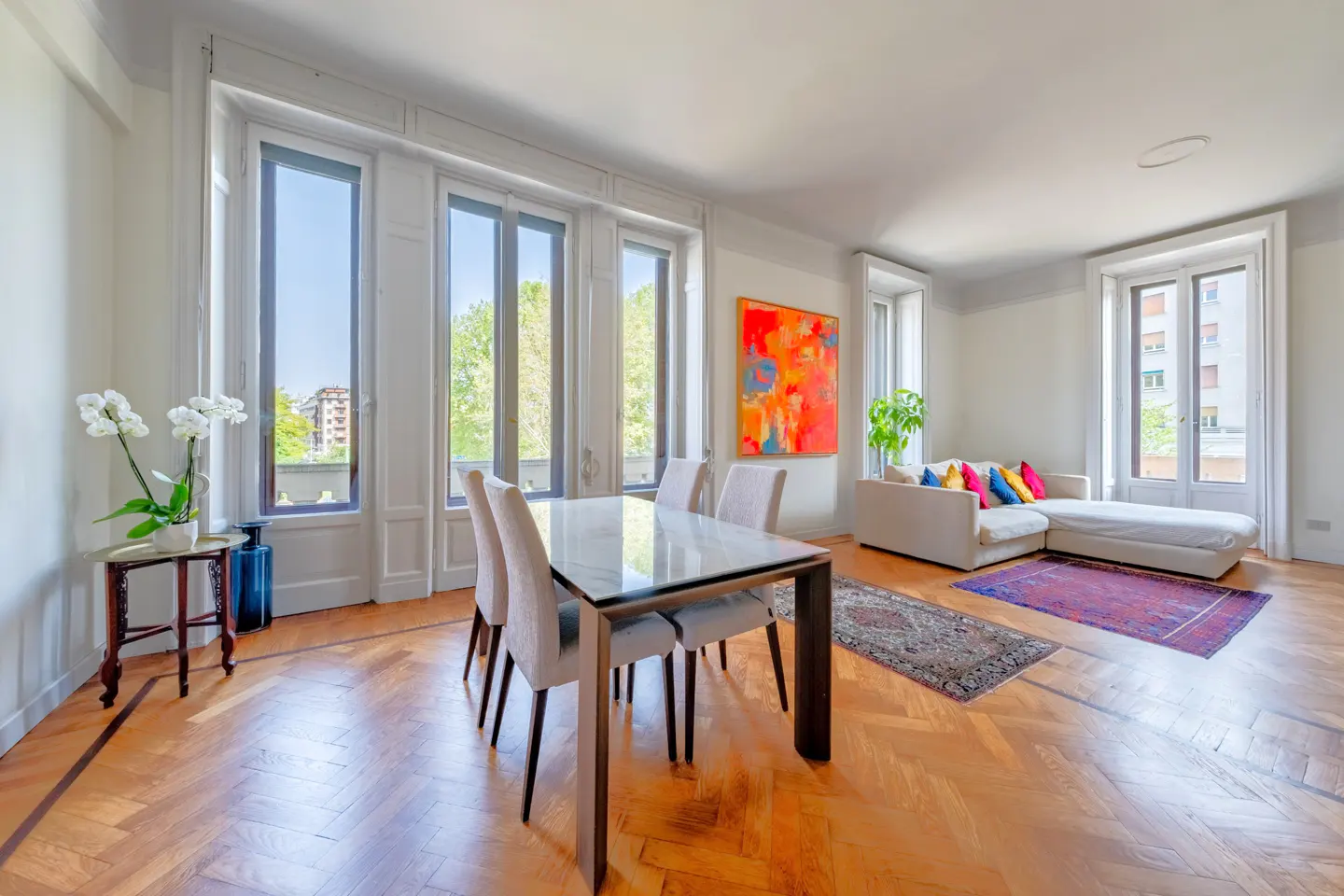 Bright, modern living room with a glass-top dining table, white sofa with colorful pillows, and large windows. Hardwood floors and an abstract painting add warmth.