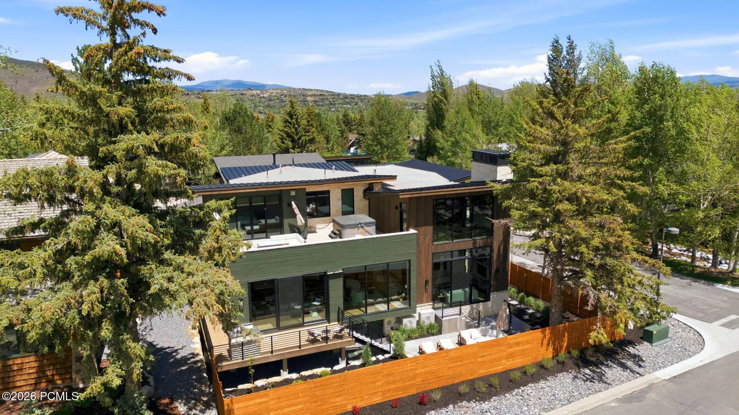 Modern home with green and brown siding, large windows, a deck, and a hot tub on the roof, surrounded by trees.