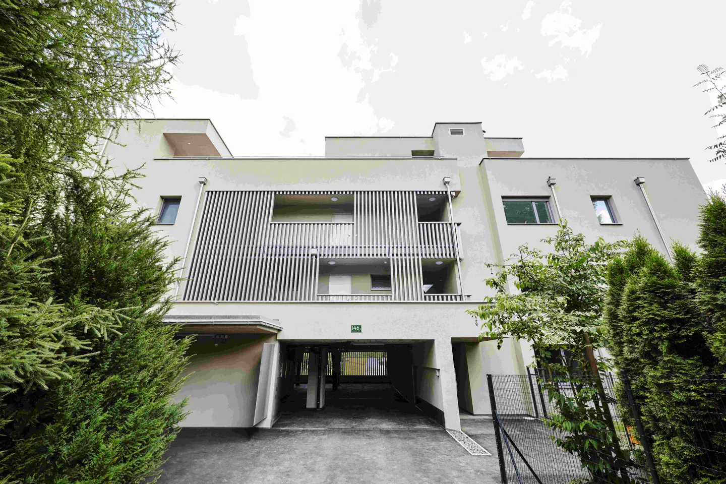 Modern light-colored apartment building with slatted balconies and a ground-level parking area, framed by green trees.